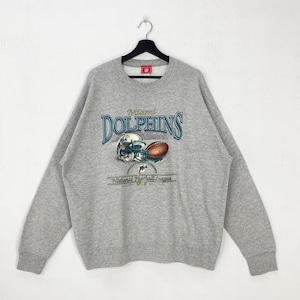 Puede incluir: Sudadera gris con el logotipo de los Miami Dolphins. El diseño incluye el nombre del equipo en escritura turquesa, un casco de fútbol americano y un balón de fútbol. Las palabras "National Football League" están impresas debajo.