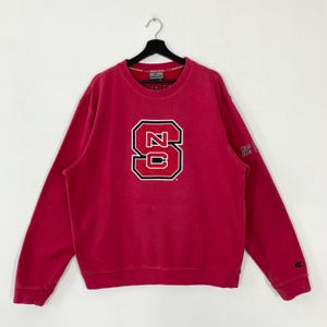 K&ouml;nnte beinhalten: Ein rotes Sweatshirt mit einem gro&szlig;en, wei&szlig; umrandeten "NC State"-Logo auf der Vorderseite. Das lang&auml;rmlige Oberteil hat einen Rundhalsausschnitt und einen kleinen "NC State"-Text auf dem rechten &Auml;rmel. Das Sweatshirt h&auml;ngt an einem schwarzen Kleiderb&uuml;gel.