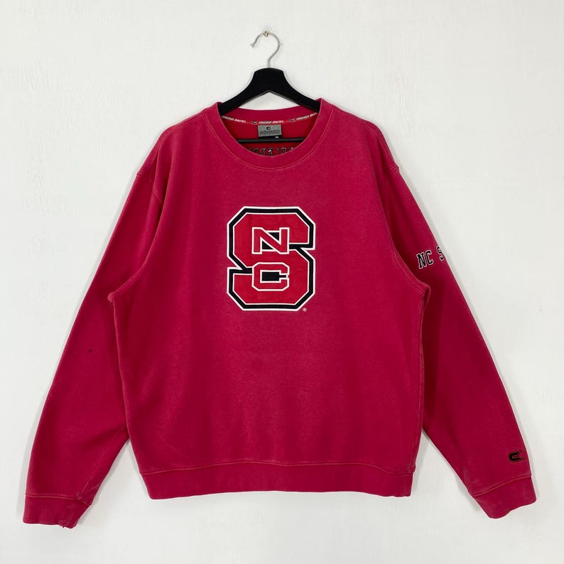K&ouml;nnte beinhalten: Ein rotes Sweatshirt mit einem gro&szlig;en, wei&szlig; umrandeten "NC State"-Logo auf der Vorderseite. Das lang&auml;rmlige Oberteil hat einen Rundhalsausschnitt und einen kleinen "NC State"-Text auf dem rechten &Auml;rmel. Das Sweatshirt h&auml;ngt an einem schwarzen Kleiderb&uuml;gel.
