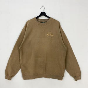 Puede incluir: Una sudadera de color marrón con cuello en V y mangas largas. Un pequeño emblema bordado está en la parte superior izquierda del pecho. La sudadera está colgada de una percha negra sobre un fondo blanco.
