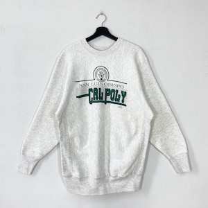 Könnte beinhalten: Ein hellgraues Sweatshirt mit dem Text "SAN LUIS OBISPO CAL POLY" in Grün und Weiß. Das Sweatshirt hängt an einem schwarzen Kleiderbügel vor einem weißen Hintergrund. Es hat lange Ärmel und einen Rundhalsausschnitt.