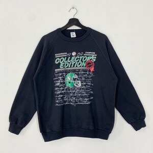 May include: A black sweatshirt with a green and white football helmet graphic. The text "ROUGHRIDERS 1989 CHAMPIONS COLLECTOR'S EDITION" is printed on the front. The design includes a red official seal and a background of signatures.