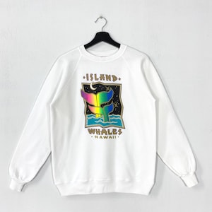 Peut inclure: Sweat-shirt blanc avec un motif coloré de queue de baleine, ciel étoilé et vagues océaniques. Le texte indique "ISLAND WHALES HAWAII". Le design présente des couleurs arc-en-ciel et des accents dorés. Le sweat-shirt est suspendu à un cintre noir.