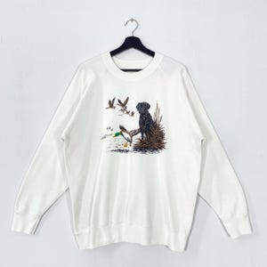 May include: White long-sleeve sweatshirt featuring a graphic of a black Labrador Retriever sitting near a duck and reeds. Several ducks are flying in the background. The sweatshirt is hanging on a black hanger.