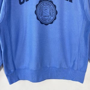 Peut inclure: Un sweat-shirt bleu clair avec un &eacute;cusson circulaire portant l'inscription "UNIVERSITY OF NORTH CAROLINA CHAPEL HILL" et le mot "LIBERTAS". Le sweat-shirt a un col rond et des manches longues.