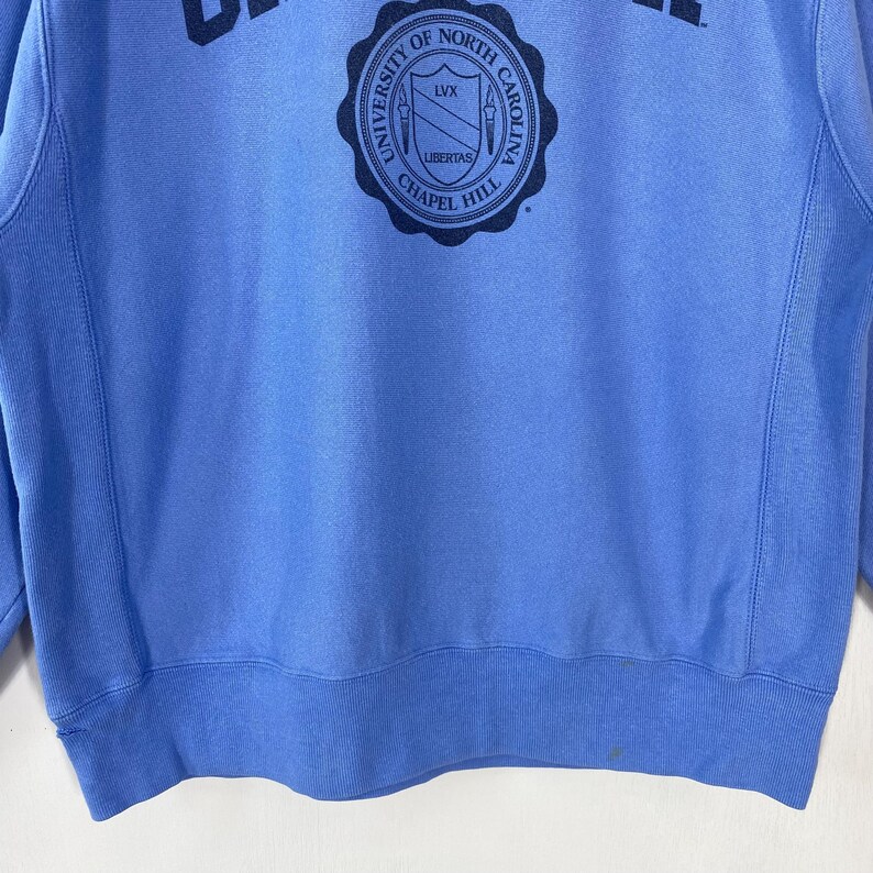 Peut inclure: Un sweat-shirt bleu clair avec un &eacute;cusson circulaire portant l'inscription "UNIVERSITY OF NORTH CAROLINA CHAPEL HILL" et le mot "LIBERTAS". Le sweat-shirt a un col rond et des manches longues.