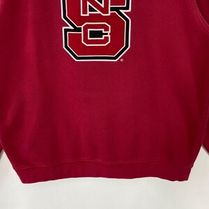 K&ouml;nnte beinhalten: Ein rotes Sweatshirt mit einer gro&szlig;en wei&szlig;en und schwarzen Grafik des Buchstabens "S" auf der Vorderseite. Die Grafik ist schwarz umrandet und hat eine rechteckige Form. Das Sweatshirt ist aus weichem Material.