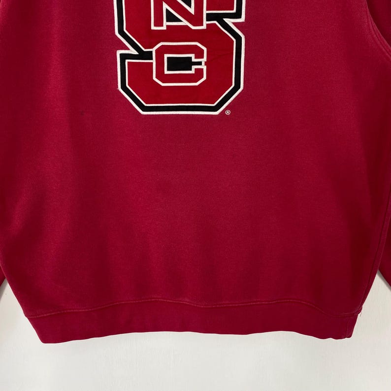 K&ouml;nnte beinhalten: Ein rotes Sweatshirt mit einer gro&szlig;en wei&szlig;en und schwarzen Grafik des Buchstabens "S" auf der Vorderseite. Die Grafik ist schwarz umrandet und hat eine rechteckige Form. Das Sweatshirt ist aus weichem Material.