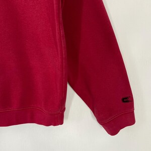 K&ouml;nnte beinhalten: Nahaufnahme eines roten Sweatshirt-&Auml;rmels. Ein schwarzes Logo ist in der N&auml;he des B&uuml;ndchens zu sehen. Der Stoff scheint weich und geb&uuml;rstet zu sein. Das Sweatshirt ist einfarbig mit einem schlichten Design.
