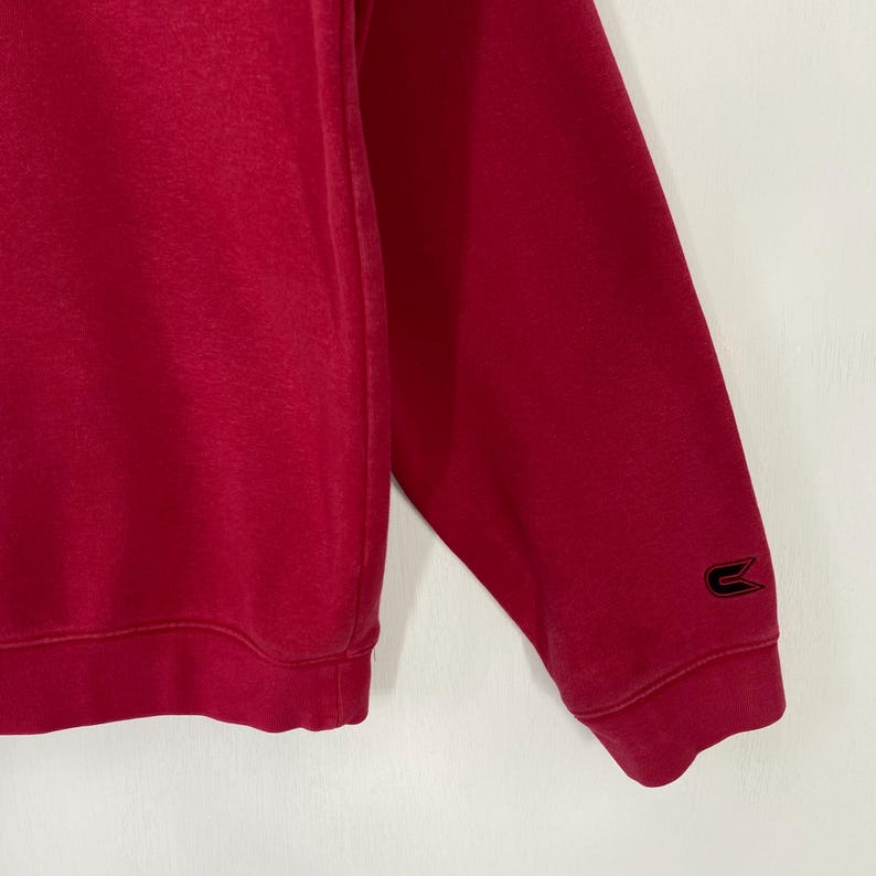 K&ouml;nnte beinhalten: Nahaufnahme eines roten Sweatshirt-&Auml;rmels. Ein schwarzes Logo ist in der N&auml;he des B&uuml;ndchens zu sehen. Der Stoff scheint weich und geb&uuml;rstet zu sein. Das Sweatshirt ist einfarbig mit einem schlichten Design.