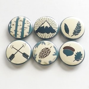 May include: Six round, blue-rimmed buttons with various nature-themed designs. The designs include a mountain, pine cones, feathers, arrows, birch trees, and an acorn.