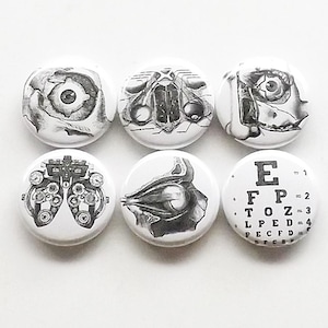 May include: Set of six black and white anatomical eye themed buttons. The buttons feature illustrations of the eye, eye muscles, and an eye chart. The eye chart reads "E, FP, TOZ, LPED, PECFD, FCZ".