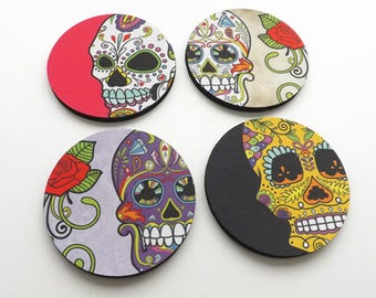 May include: Four round coasters with a Day of the Dead theme. Each coaster features a colorful sugar skull with intricate designs and floral accents. The coasters are black, red, white, and yellow.