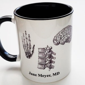 May include: White ceramic coffee mug with a black handle. The mug has a black rim and features a black and white illustration of a human skeleton hand, spine, and brain. The text "Jane Meyer, MD" is printed on the bottom of the mug.
