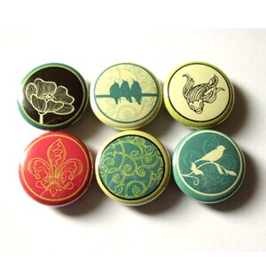 May include: Six round button pins with different designs. The designs include a flower, birds, a fish, a fleur-de-lis, and two different patterns.