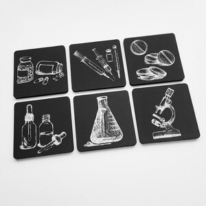 May include: Six black square coasters with white illustrations of science-related items. The coasters feature images of pills, a syringe, a beaker, a dropper bottle, a microscope, and a flask.