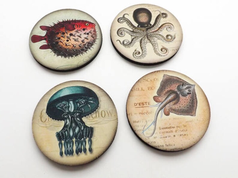 May include: Four round wooden coasters with vintage illustrations of sea creatures. The coasters feature a pufferfish, an octopus, a jellyfish, and a ray. The coasters are printed with a light brown background and black text.