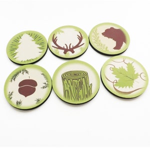 May include: Six round green and white coasters with different nature-themed designs. The designs include a tree, a deer, a bear, a tree stump, an acorn, and a leaf.