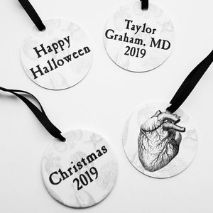 May include: Three round white ornaments with black ribbon hangers. The ornaments have black text on a white background. The text on the ornaments reads "Happy Halloween", "Christmas 2019", and "Taylor Graham, MD 2019".