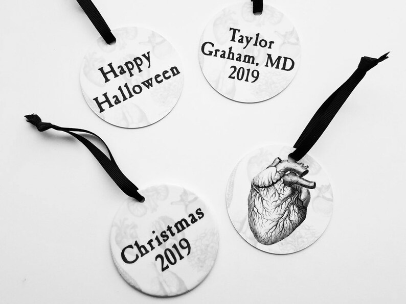 May include: Three round white ornaments with black ribbon hangers. The ornaments have black text on a white background. The text on the ornaments reads "Happy Halloween", "Christmas 2019", and "Taylor Graham, MD 2019".