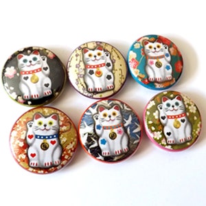 May include: Six round, colorful buttons with a white cat with a raised paw, a bell around its neck, and a black heart on its chest. The cat is on a patterned background. The buttons are approximately 2 inches in diameter.