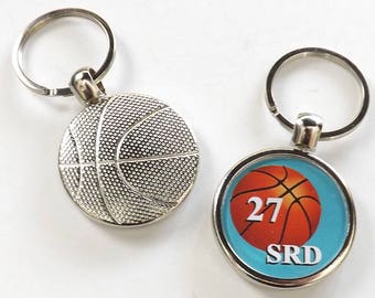 May include: Two silver basketball keychain charms. One charm features a silver basketball with a textured surface. The other charm features a round, blue background with a basketball graphic, the number 27, and the text "SRD".