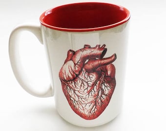 May include: White ceramic mug with a red interior and handle. The mug features a detailed illustration of a human heart in shades of red and brown. The mug is a standard size for coffee or tea.