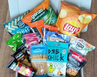 Può includere: Una varietà di snack in una scatola di cartone. Gli snack includono patatine fritte, cracker, pretzel e anelli di cipolla disidratati. La scatola è piena di diversi marchi, tra cui Lay's, Flint e Gonzo.