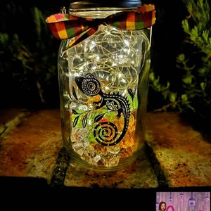 May include: A glass jar with a black and white lizard design, filled with clear crystals and fairy lights. The jar is decorated with a multi-colored ribbon and has a silver lid. The jar is sitting on a brown brick surface.