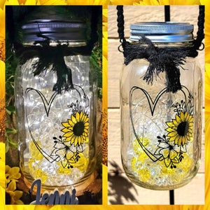 May include: A mason jar with a black ribbon and a heart-shaped design with sunflowers and other flowers. The jar has a solar-powered light inside and the word "Jenni" is written on a wooden plaque below the jar.