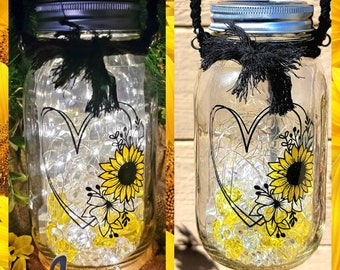 May include: A mason jar with a black ribbon and a heart-shaped design with sunflowers and other flowers. The jar has a solar-powered light inside and the word "Jenni" is written on a wooden plaque below the jar.