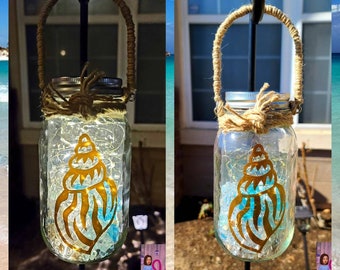 May include: Two glass jars with a seashell design and string handles. The jars are filled with white lights and blue and white glass beads. The jars are hanging from a metal hook.
