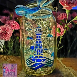 May include: A glass mason jar with a blue ribbon and a lighthouse design. The jar is filled with white lights and blue and white seashells. The jar is sitting on a wooden surface.