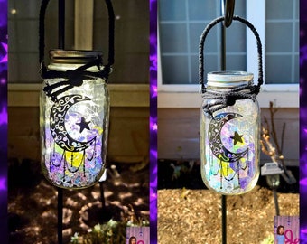 May include: A mason jar with a crescent moon and star design is hanging from a metal hook. The jar is filled with iridescent glass beads and has a solar powered light inside. The jar is hanging in a garden setting.