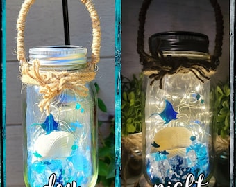 May include: A mason jar with a rope handle filled with blue and white sea glass, a seashell, and a blue stingray. The jar is lit up with white fairy lights and the text "day" is written on the bottom of the jar. The jar is shown next to a similar jar with the text "night" on the bottom.