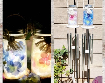 May include: Two glass jars with white lids and rope handles, filled with pressed flowers and lit from within. The jars are on a wooden surface. Two metal wind chimes with silver tubes hang from a black metal frame. The wind chimes are on a wooden surface.