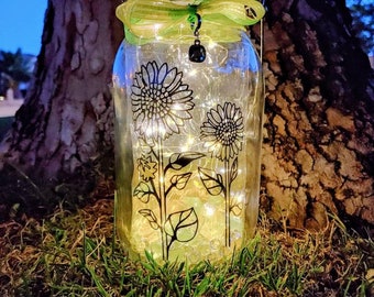 May include: A mason jar with a green ribbon and a black charm, decorated with black and white sunflower designs. The jar is filled with clear glass beads and white fairy lights.