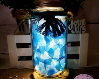 May include: A mason jar with a blue and white floral design, a blue ribbon, and a wooden base. The jar is lit from within, creating a warm glow. The jar is labeled "Jenni Jars Original & Authentic".
