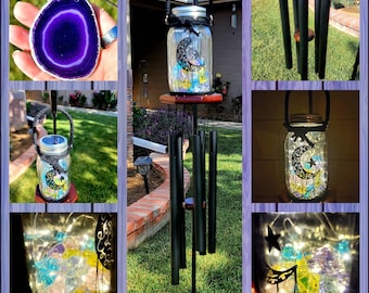May include: A solar-powered mason jar wind chime with a crescent moon and stars design. The jar is filled with iridescent glass beads and lights up at night. The wind chime is hanging from a black metal stand with four tubes.