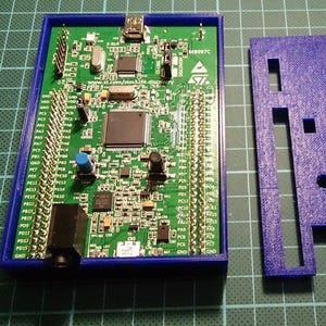 Pode incluir: Uma placa de circuito verde com vários componentes eletrônicos, incluindo chips, conectores e uma porta USB, está alojada em uma estrutura retangular azul. Uma tampa azul separada com recortes está ao lado. Em uma grade verde.