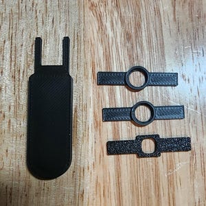 May include: A collection of black 3D-printed parts, including a rectangular piece with a handle and three smaller pieces with circular and rectangular openings. The items are arranged on a wooden surface.