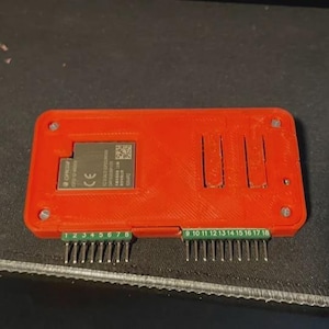 May include: A red electronic component with a rectangular form. It has a small, embedded circuit board and two rows of pins labelled with numbers. The component has ventilation slots and is mounted on a dark surface.