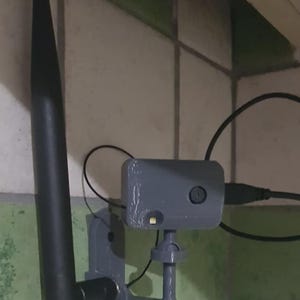 Może przedstawiać: Szare, drukowane w 3D urządzenie z czarną anteną i małym światłem. Urządzenie jest zamontowane na ścianie i podłączone do czarnego kabla. Urządzenie wydaje się być czujnikiem lub małą kamerą.