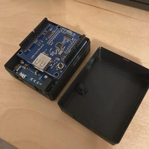 Puede incluir: Una caja de dispositivo electrónico negra con una placa de circuito azul en el interior. La placa de circuito tiene varios componentes, incluido un puerto USB y el texto "USB Host Shield". La caja está abierta, revelando los componentes internos.