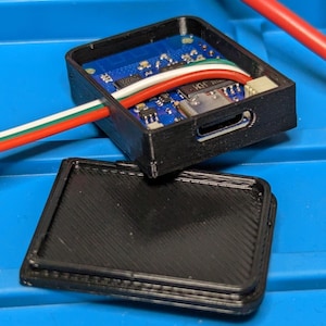 May include: A black 3D-printed rectangular enclosure with an open top, revealing a blue circuit board with a USB-C port and multi-coloured wiring. A matching black rectangular lid with a textured diagonal surface finish lies separately in front.