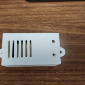 May include: A small, white rectangular electronic component with a series of vertical slots on one side and two small holes on the other. It has two small, rounded loops on the sides.