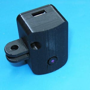 May include: A black 3D printed camera mount with a GoPro-style attachment and a small lens opening. The mount has a slot for a micro USB cable.