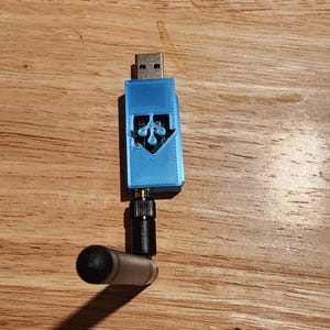 Può includere: Un dispositivo USB blu con un'antenna nera. Il dispositivo ha un logo sulla parte anteriore ed è collegato a un'antenna nera con un'estremità arrotondata. Il dispositivo è su una superficie di legno.