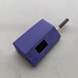 Pode incluir: Um dispositivo eletrónico retangular roxo com acabamento mate, apresentando um pequeno ecrã retangular preto, dois ícones circulares gravados que se assemelham a termómetros e uma antena preta no canto superior direito.