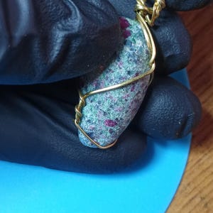 May include: A pendant featuring a rough, oval-shaped stone with a speckled green and grey texture and embedded deep pink inclusions. The stone is secured in a gold-colored metal wire wrap that forms a loop at the top for a chain or cord attachment.